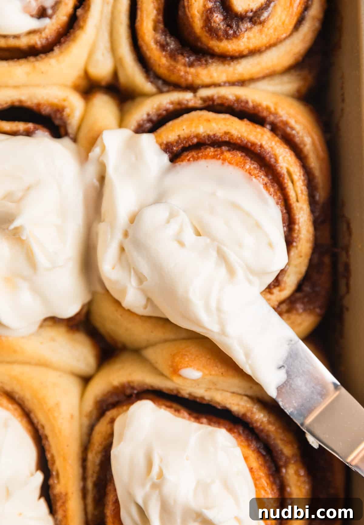 Fluffy Buttermilk Cinnamon Rolls from Scratch 2 Cinnamon rolls in baking pan with icing being spread over top with offset spatula.