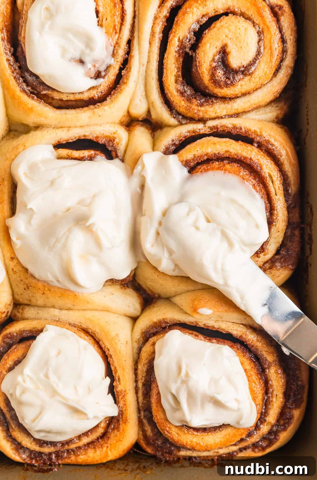 Fluffy Buttermilk Cinnamon Rolls from Scratch 21 Frosting being spread on freshly baked cinnamon rolls in pan.