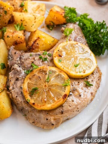 Lemon pepper pork chop on white plate with lemon slices and freshly chopped parsley.
