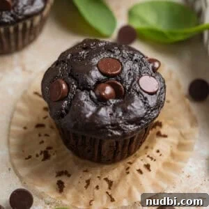 Chocolate spinach muffin sitting on open muffin liner with chocolate chips and spinach surrounding.