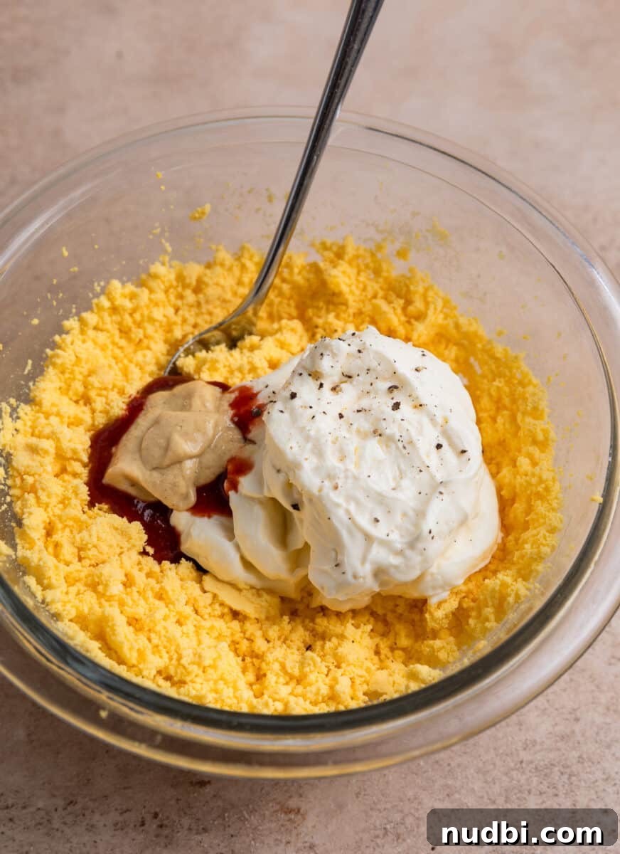 Spicy Deviled Eggs Recipe with Tangy Jalapeño Kick 6 Mashed egg yolk, mayonnaise, dijon mustard and other ingredients in glass bowl with spoon.