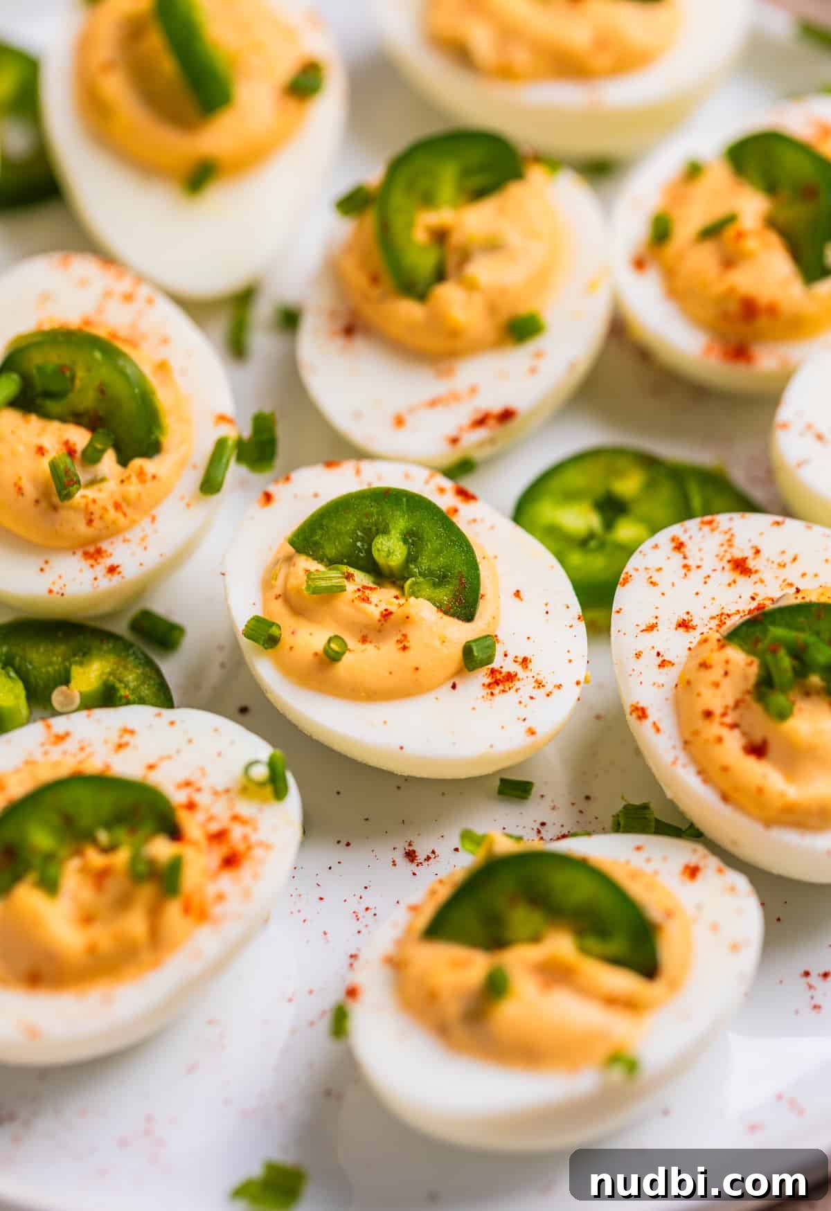 Spicy Deviled Eggs Recipe with Tangy Jalapeño Kick 10 White dish with spicy jalapeño deviled eggs garnished with chives, paprika and sliced jalapeño.