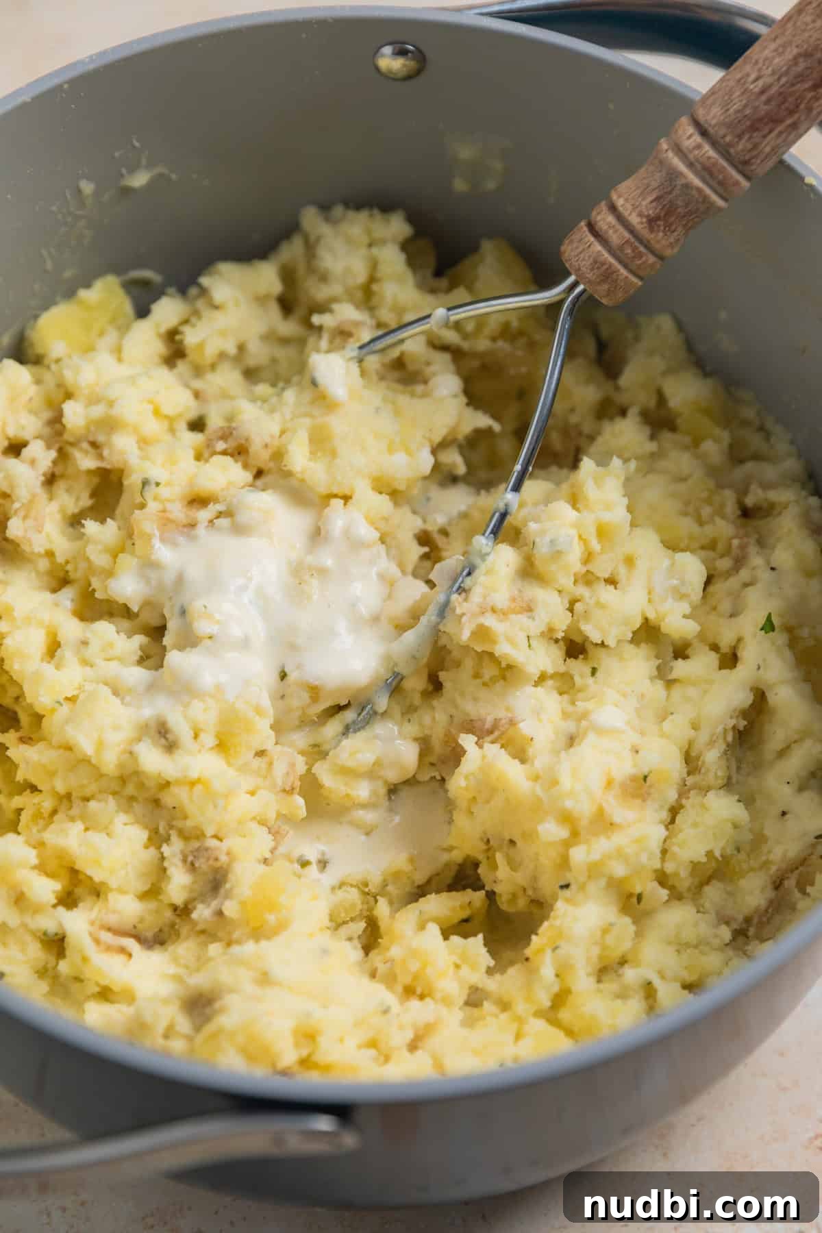 Creamy Boursin Mashed Potatoes Recipe for Garlic-Herb Flavor 6 Potatoes being mashed with potato masher and cream, cheese and butter mixture added on top.