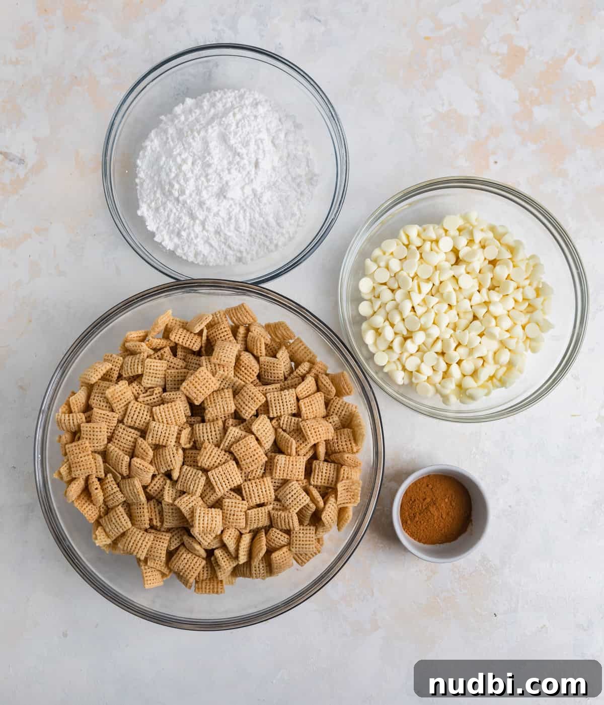 Snickerdoodle Puppy Chow Recipe — Cinnamon Sugar Snack Mix 3 Powdered sugar, white chocolate chips, cinnamon and Chex cereal on counter.