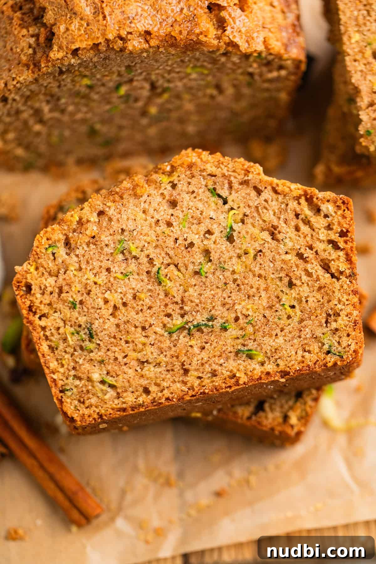 Grandma’s Zucchini Bread Recipe (Even Better) 2 Slices of zucchini bread on parchment with cinnamon sticks beside.
