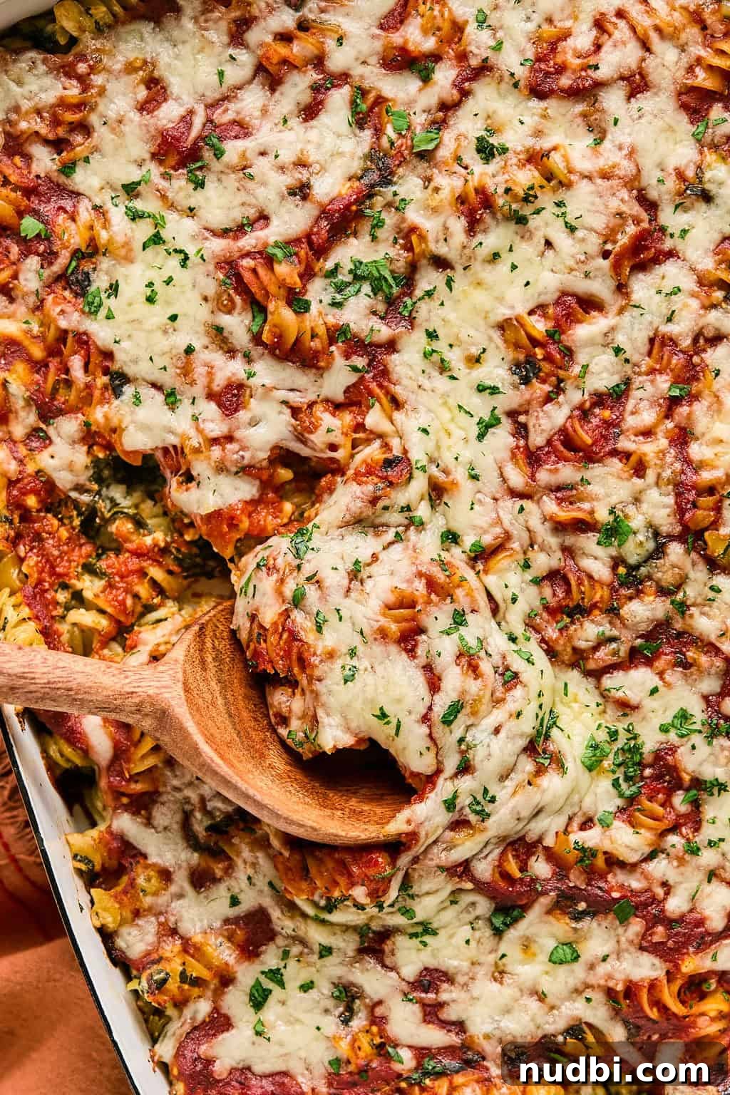 Spinach Baked Pasta with Creamy Cheese and Garlic 2 Wooden spoon scooping into baked pasta with spinach topped with chopped parsley.