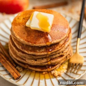 Stack of pancakes on plate with butter and maple syrup and cinnamon sticks beside.