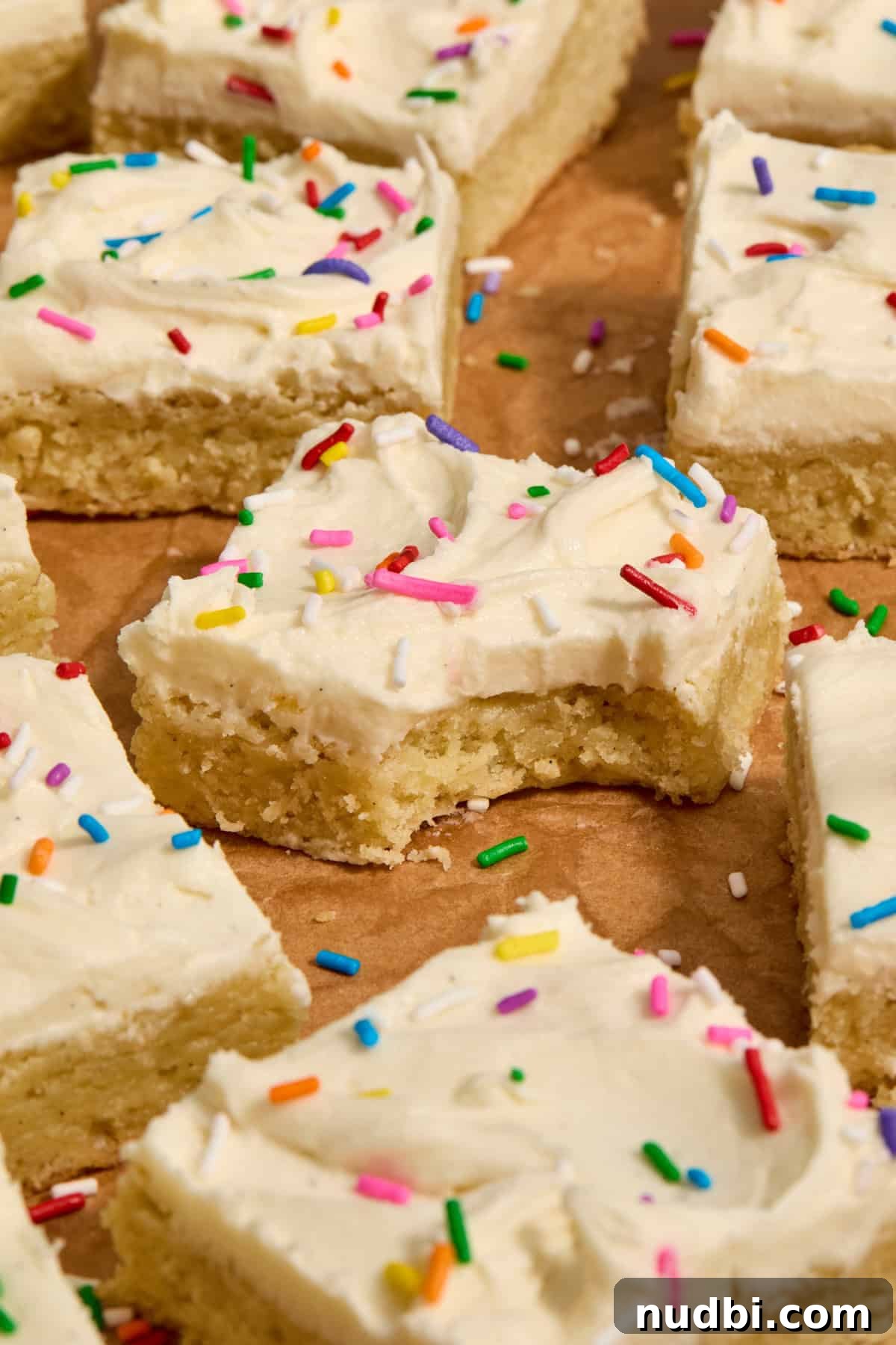 Frosted Sugar Cookie Bars Recipe — Soft, Chewy Dessert Bars 2 Vanilla frosted sugar cookie bars with rainbow sprinkles on top on parchment.