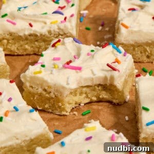 Frosted Sugar Cookie Bars Recipe — Soft, Chewy Dessert Bars 18 Vanilla bean frosted sugar cookie bars with rainbow sprinkles on parchment.
