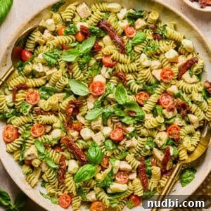 Pesto Chicken Pasta Salad with Sun-Dried Tomatoes and Basil 12 Pesto chicken pasta salad in serving bowl topped with fresh basil with serving spoon.