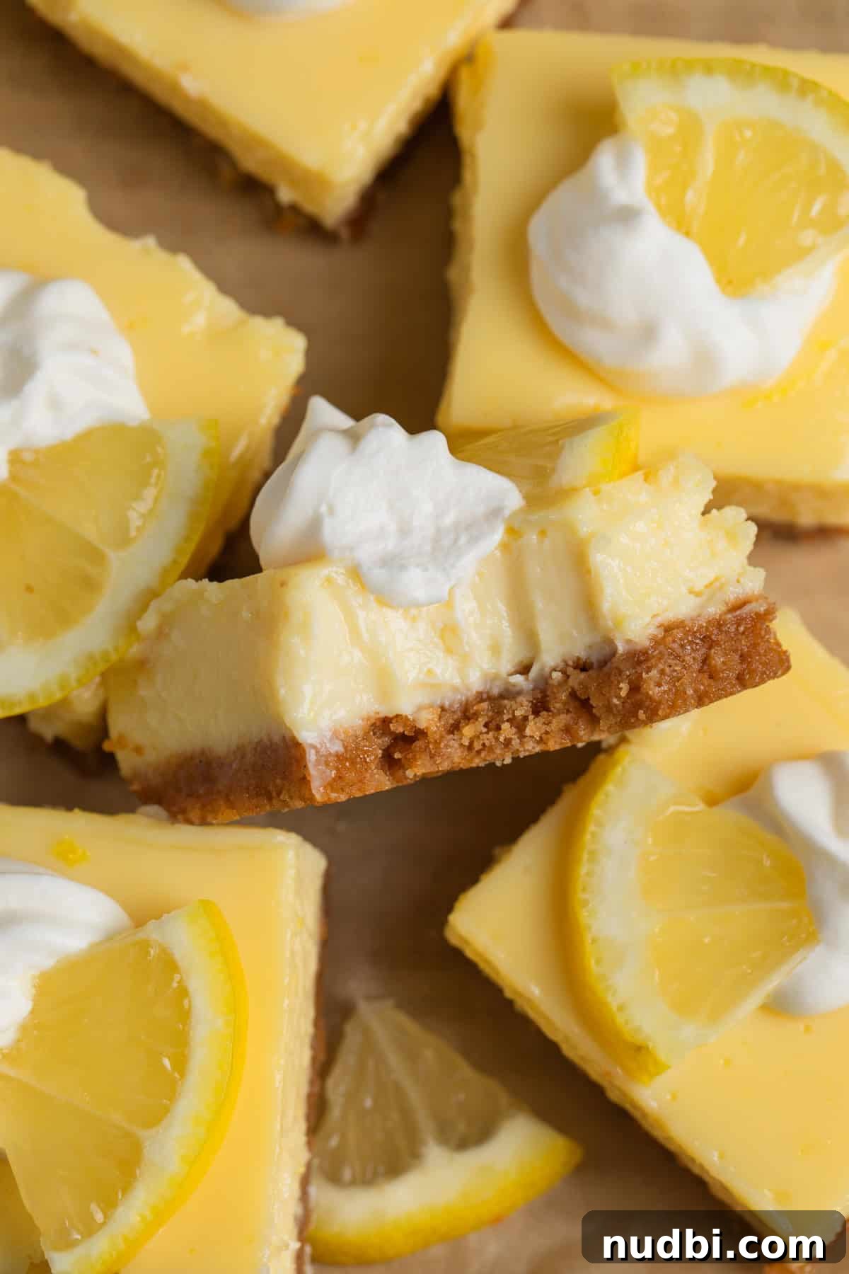 Creamy Lemon Bars with Graham Cracker Crust Recipe 2 Creamy lemon bars on parchment with bite taken out of one to show texture.
