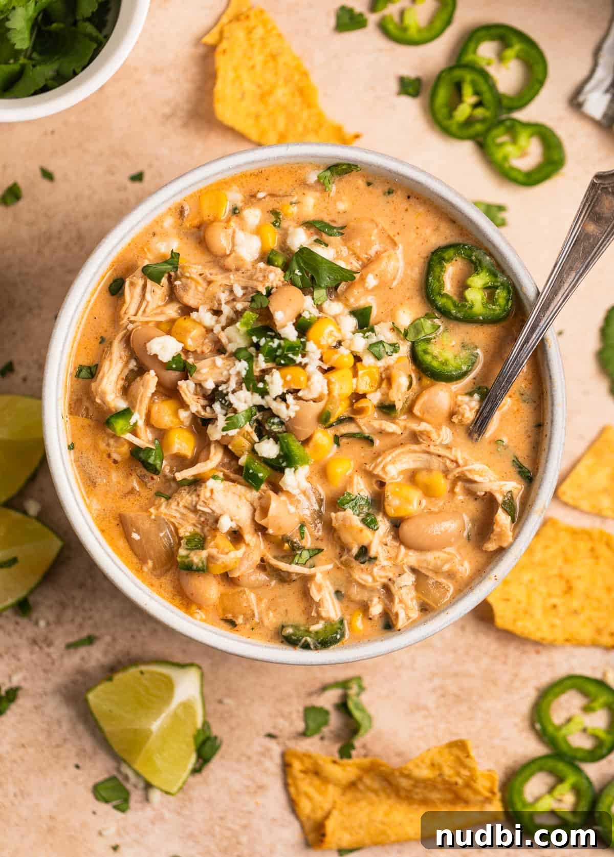 Creamy Slow Cooker White Chicken Chili Recipe 2 Slow cooker creamy white chicken chili in bowl topped with queso fresca, jalapeno, cilantro and spoon scooping into the soup and chips and limes next to it.