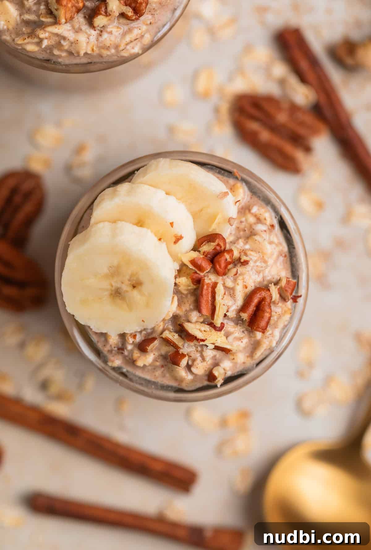 Banana Bread Overnight Oats Recipe for Creamy Breakfast Bliss 6 Glass jar with banana bread overnight oatmeal garnished with banana and pecans and cinnamon sticks, pecans and spoon surrounding.