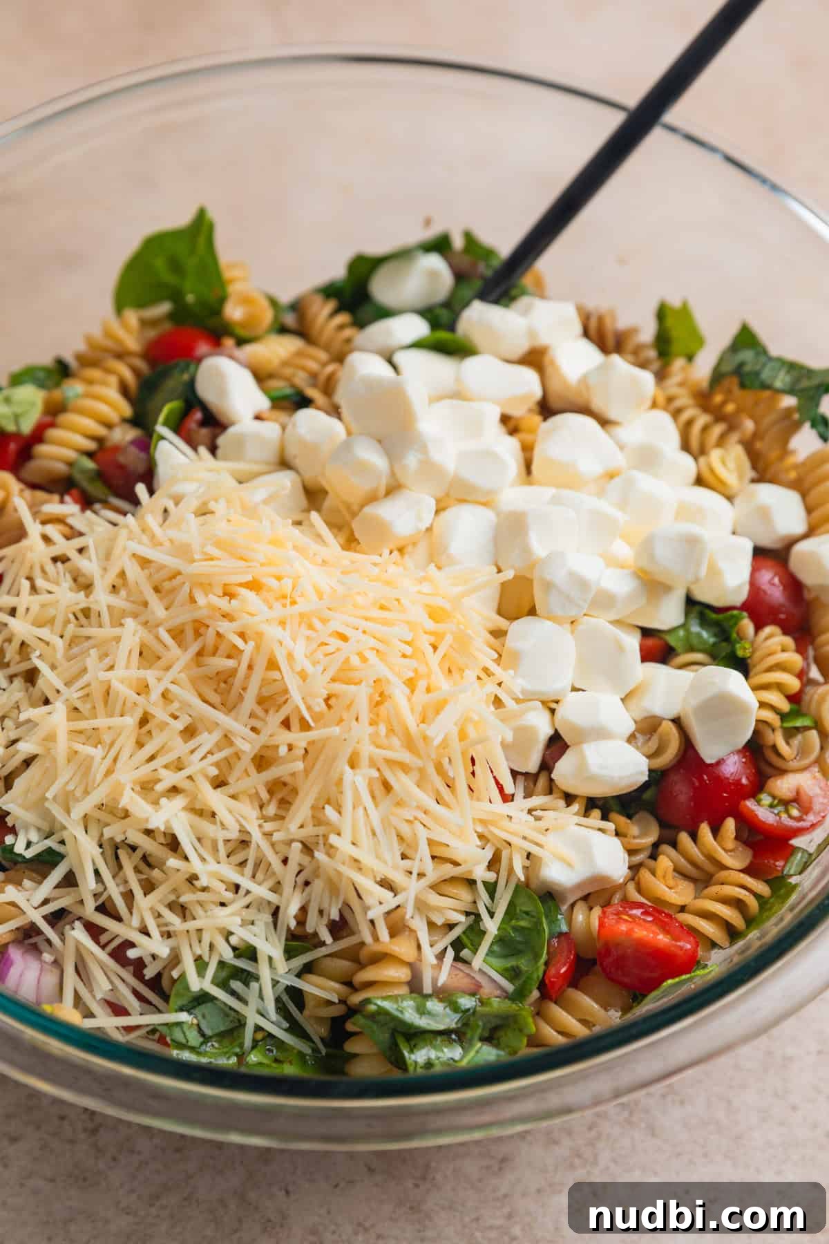 Shredded parmesan and fresh mozzarella pearls on top of rotini and vegetable mixture in bowl.