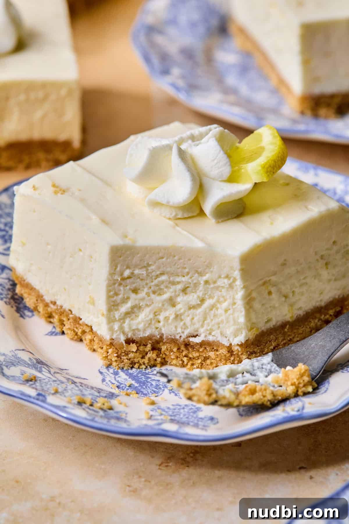 No-Bake Lemon Cheesecake Bars with Zesty Graham Crust 15 Slice of no-bake lemon cheesecake bar on plate with forkful taken out to show texture.
