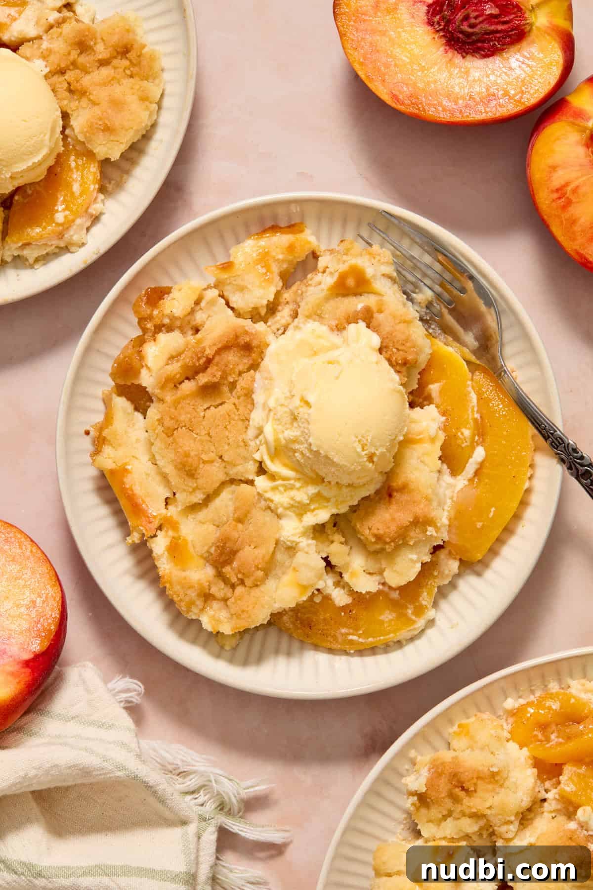 Cream Cheese Peach Cobbler with Buttery Biscuit Topping 2 Cream cheese peach cobbler with scoop of vanilla ice cream on plate with fork and peaches surrounding.
